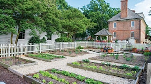 Un jardin avec des plates-bandes surélevées, une clôture en bois peinte en blanc et un bâtiment en briques doté d’une cheminée.