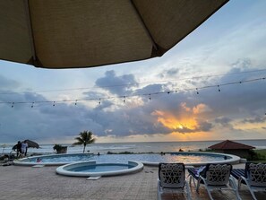 Un espace piscine comprenant plusieurs piscines, des chaises longues et une vue sur l’océan au coucher du soleil.