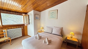 A bedroom with a wooden ceiling, a large bed with white linens, two bedside lamps, a radiator, and a window with a view of trees.