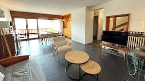 A spacious living area with a sofa, two round tables, a TV, and a balcony with a wooden railing.