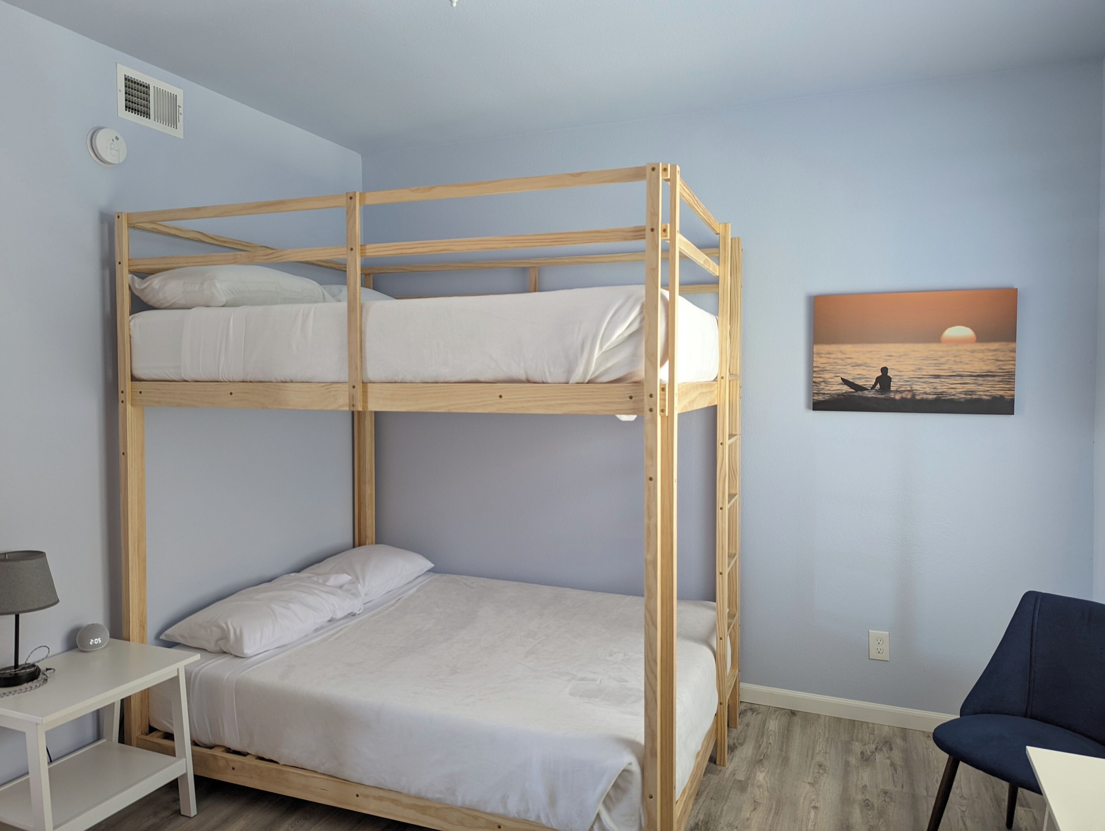 Een slaapkamer met stapelbed, nachtkastje, stoel en een schilderij van een zonsondergang boven de oceaan.