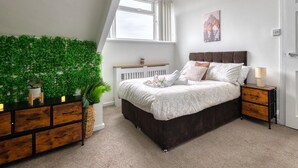 Een slaapkamer met een schuine wand, een bed met donker hoofdeinde, een nachtkastje met lamp, een hangende plant aan de muur en een raam met uitzicht op de lucht.