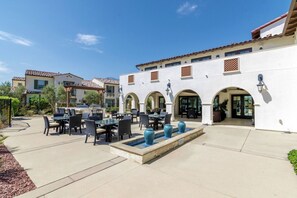 An outdoor patio area with tables and chairs, a small fountain, and a building with arched doorways.
