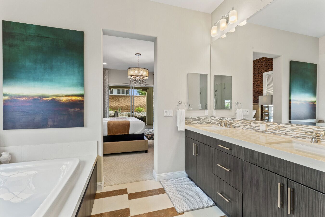 A modern bathroom with a large mirror, a bathtub, and a vanity with a sink. There is a painting on the wall and a view of a bedroom with a bed and a chandelier.