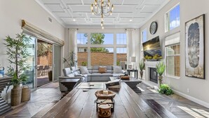 A spacious living room with a large sectional sofa, a wooden coffee table, and a fireplace. There are large windows providing natural light and a view of the outdoors. - 2BR retreat - hot tub, gym & Coachella nearby (La Quinta)