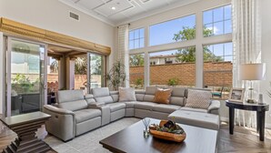 A spacious living room with a large sectional sofa, a wooden coffee table, and a view of a brick wall and greenery outside. - 2BR retreat - hot tub, gym & Coachella nearby (La Quinta)