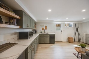 A modern kitchen with grey cabinets, a white countertop, and a microwave. There is a wooden shelf with books and decorative items. A wicker basket and a small table with a plant are visible in the living area. - Cozy Garden Suite by Sloan's Lake and Tennyson! (Wheat Ridge)
