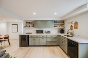 A modern kitchen with light green cabinets, a built-in microwave, toaster, and dishwasher. There are wooden shelves above the counter and a welcome sign on the wall. - Cozy Garden Suite by Sloan's Lake and Tennyson! (Wheat Ridge)