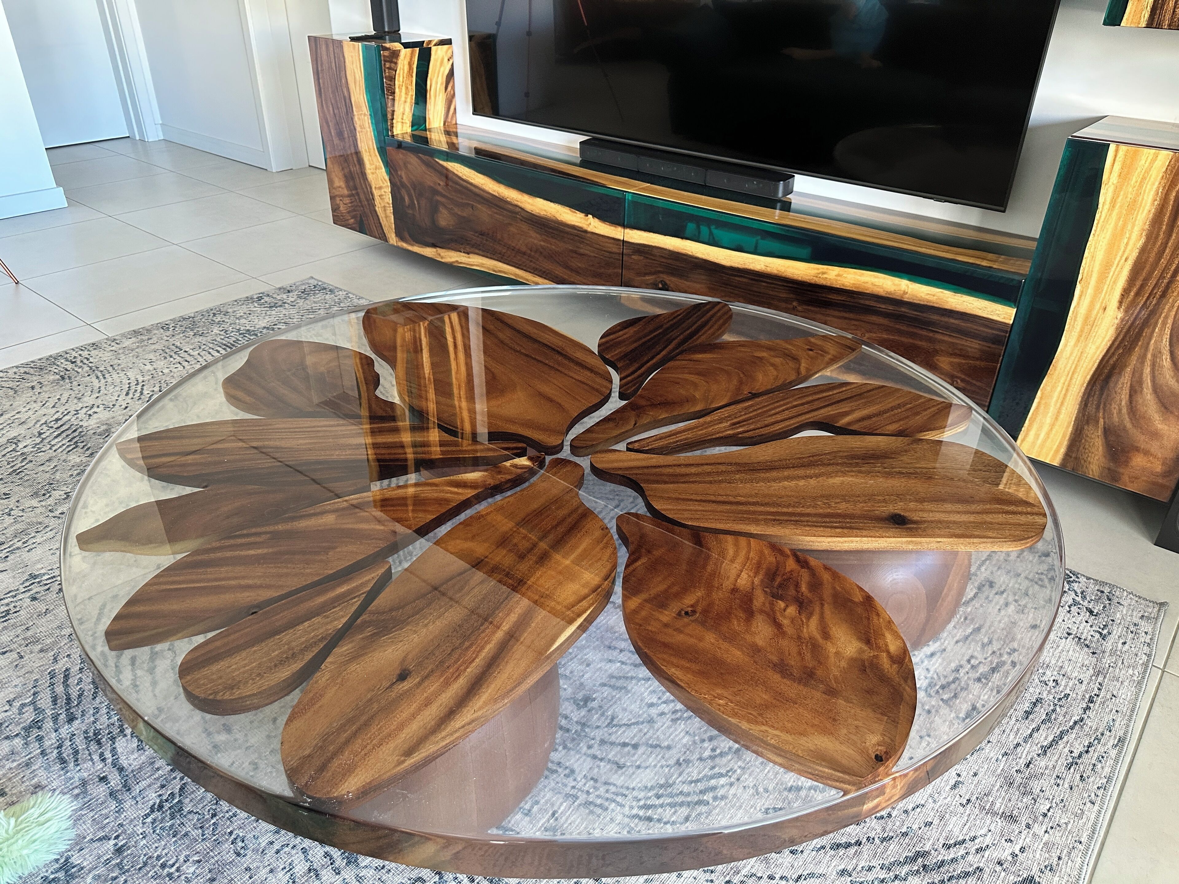 Une table basse ronde en verre, ornée de motifs en forme de feuilles en bois, un meuble TV moderne avec des éléments en bois et en verre, et un tapis à motifs.