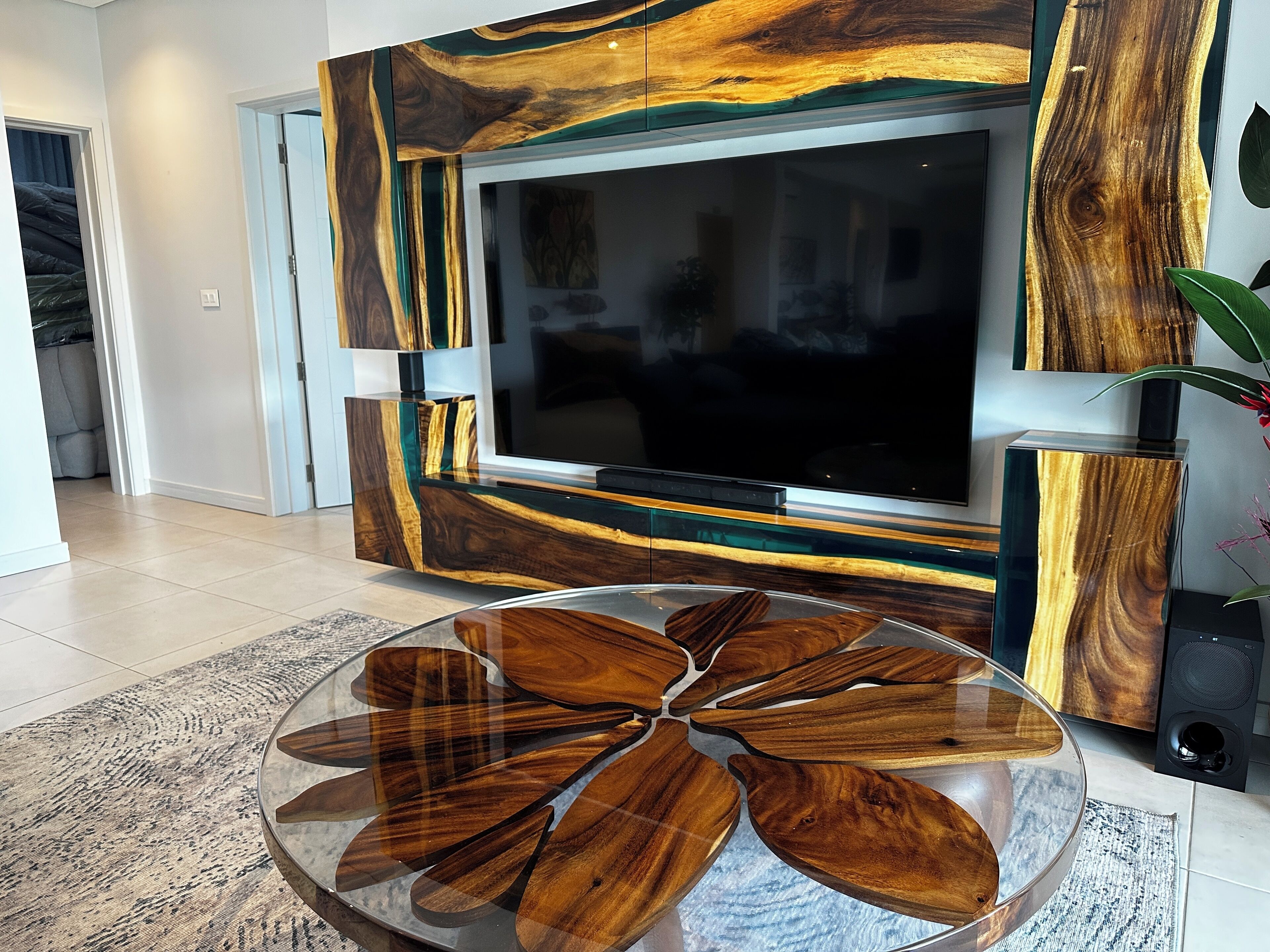 Una sala de estar moderna con un televisor de pantalla plana grande montado en una pared con diseño de vetas de madera. Hay una mesa redonda de centro con un motivo de hojas de madera frente al televisor.