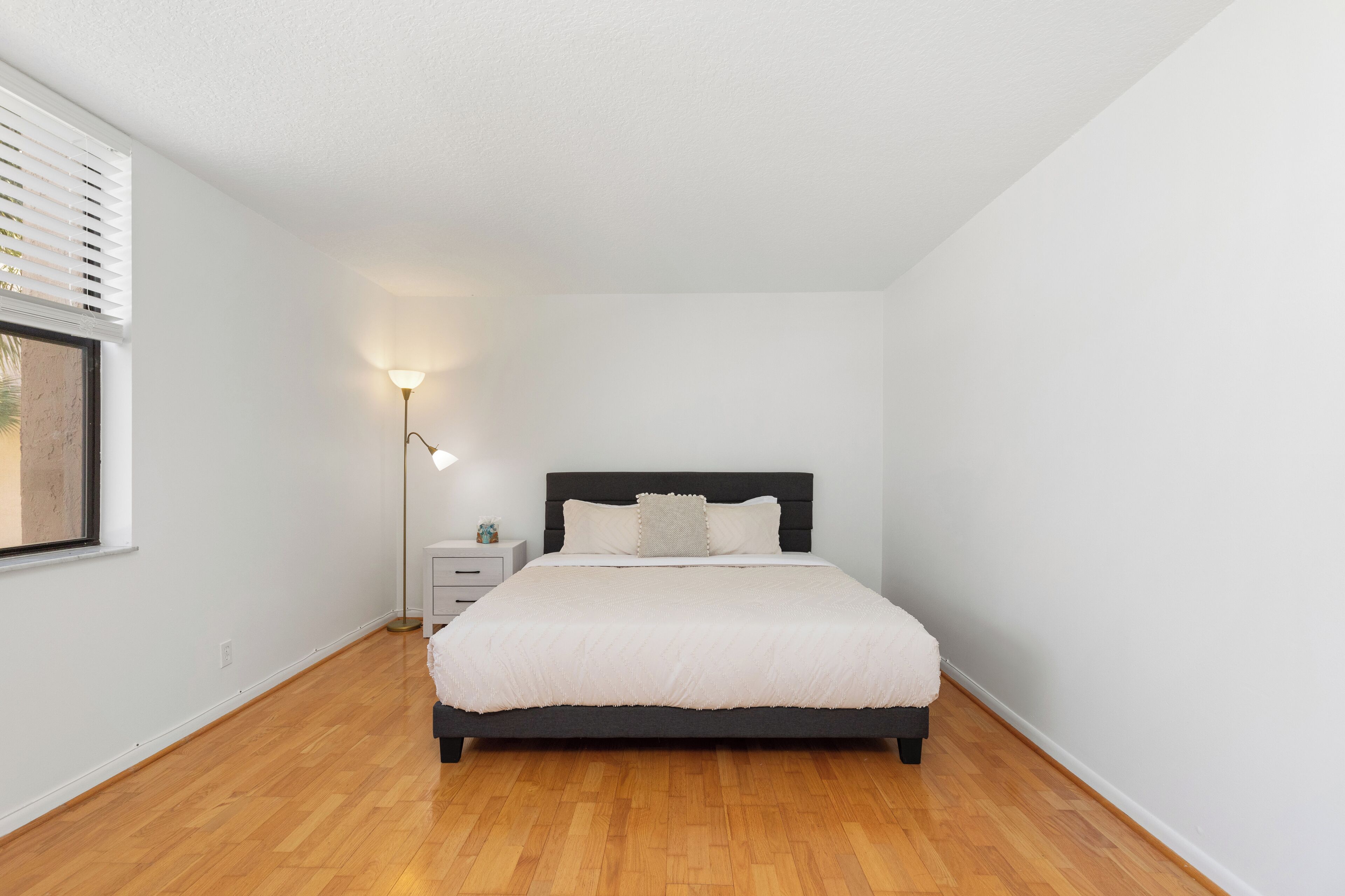 A bedroom with a bed, bedside table, lamp, and a window with blinds.