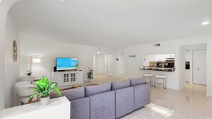 Una sala de estar moderna con un sofá gris, un televisor y una pequeña cocina al fondo.