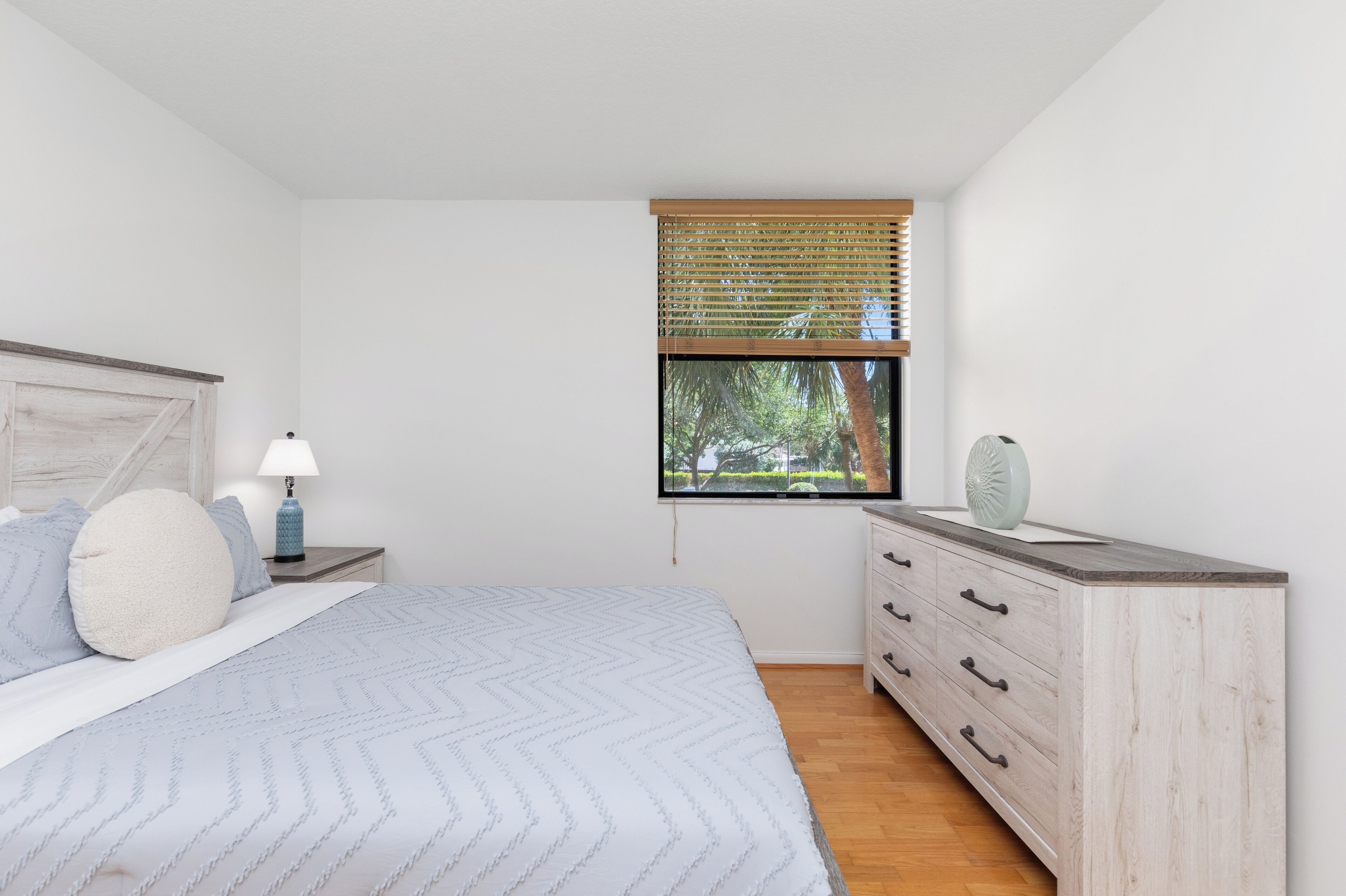 Ein Schlafzimmer mit Bett, Nachttisch, Kommode, einem Fenster mit Jalousien und Blick auf Bäume.