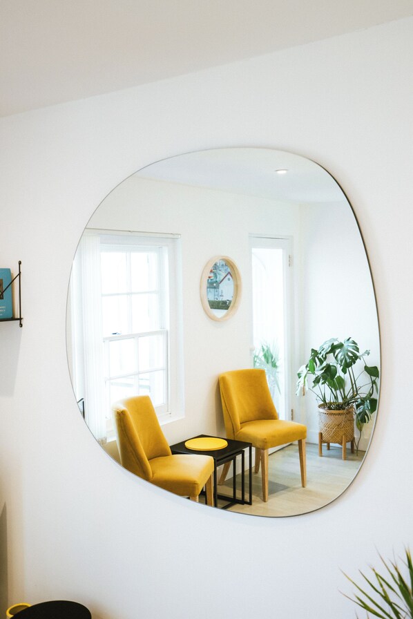 A room with a large round mirror on the wall, two yellow chairs, a small table, and a potted plant. - The Old Captains Lookout (Falmouth)