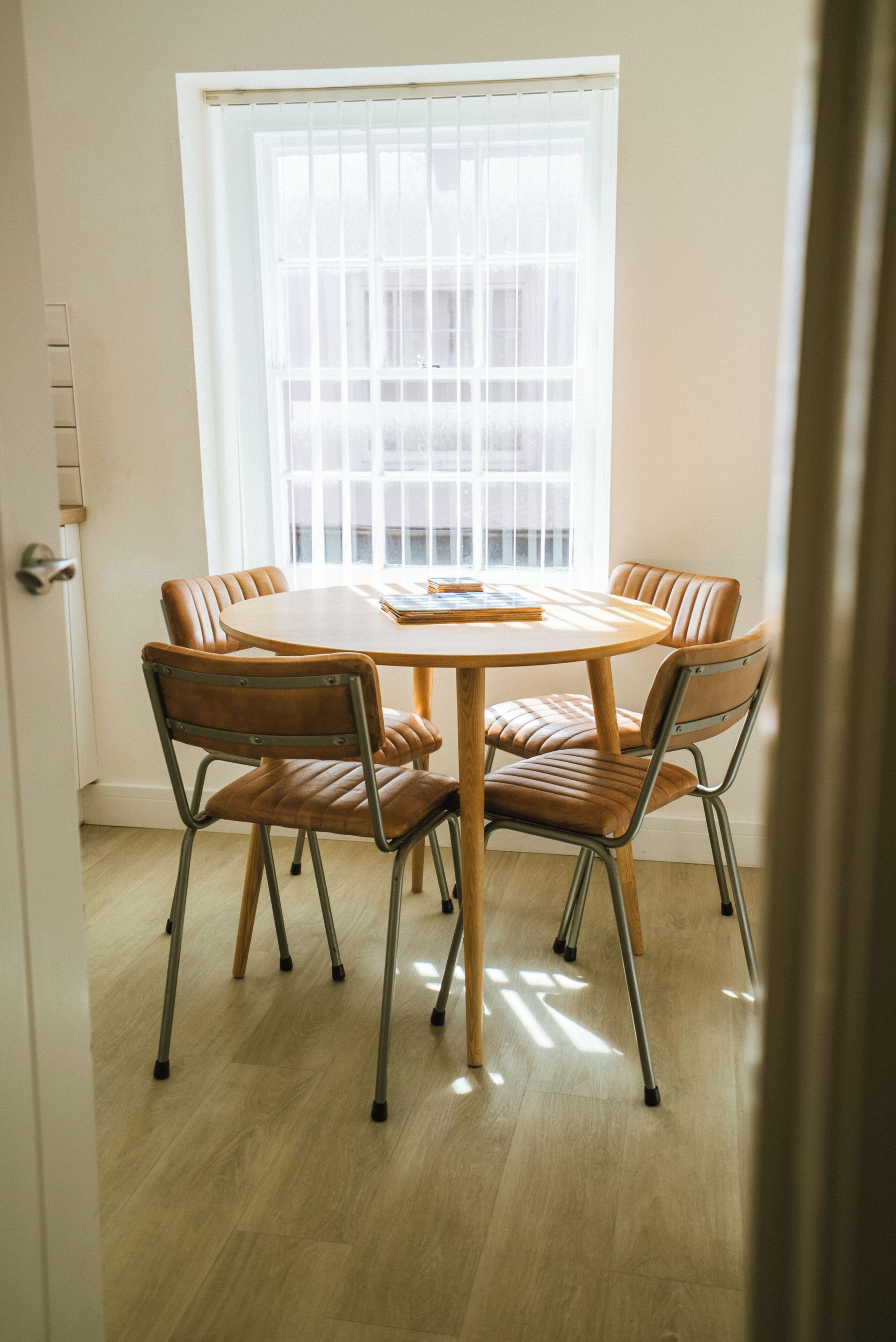 A dining area with a round table and six chairs, a window with blinds, and a door with a handle.