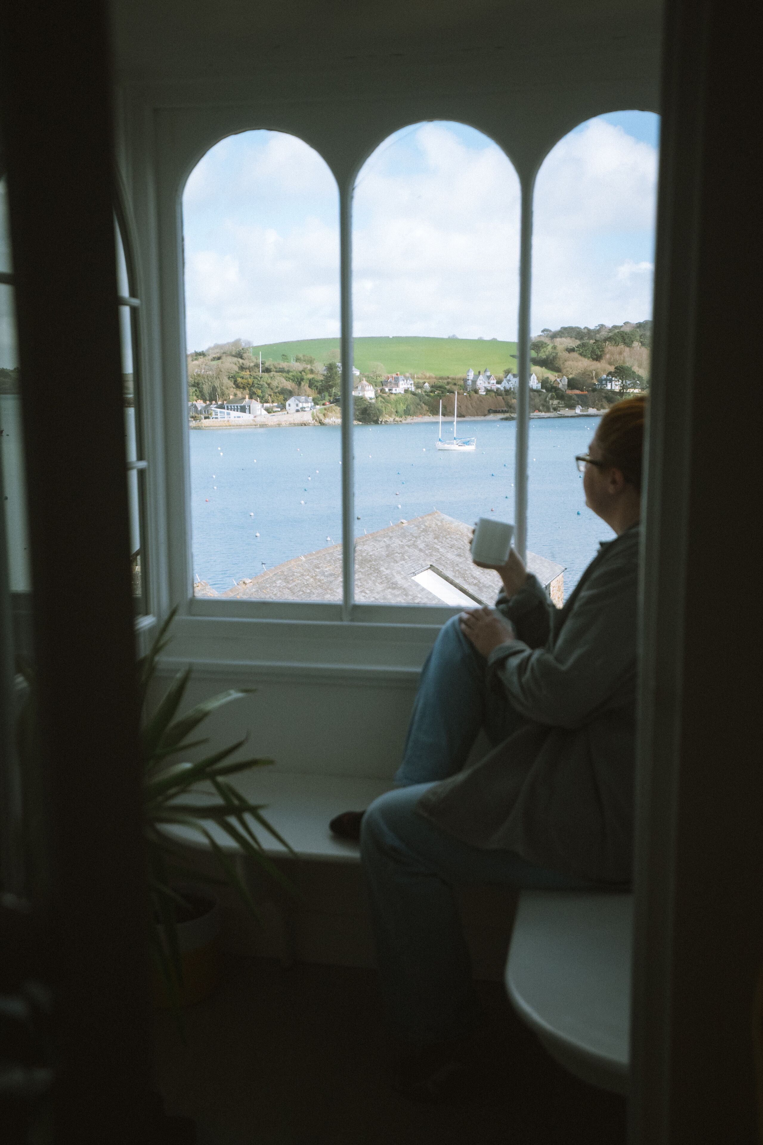 A person sitting by a window with a view of a body of water and a distant shoreline.
