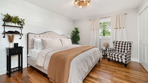 A bedroom with a bed, a chair, a bedside table, a shelf with plants, and a window with curtains. - At Home in Acworth! (Acworth)