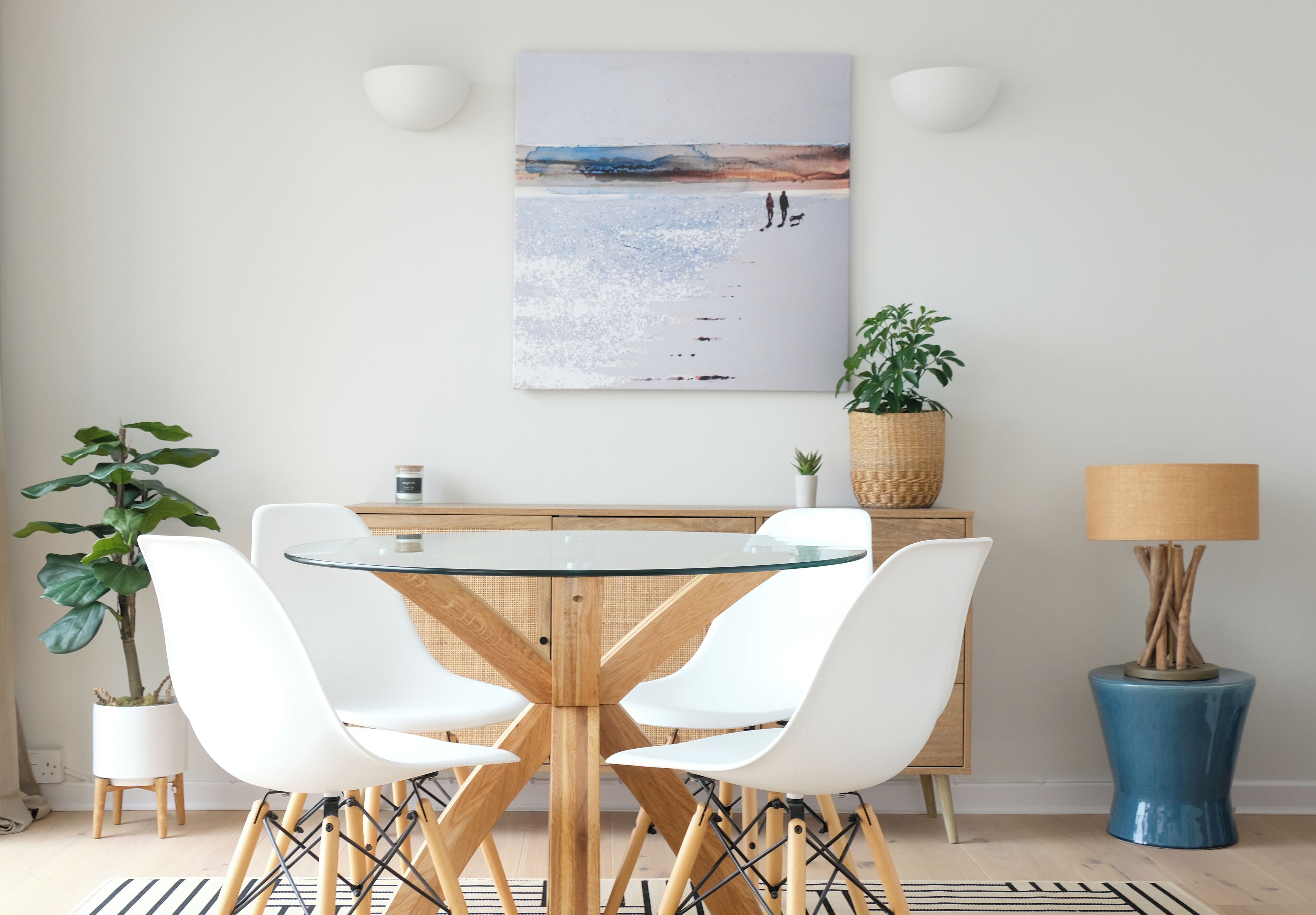 A dining area with a round glass table, white chairs, a wooden cabinet, a potted plant, and a lamp.