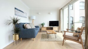 En moderne stue med en blå sofa, et træbord og et fjernsyn på et træstel. Der er et stort vindue med gardiner, en plante og en gulvlampe.