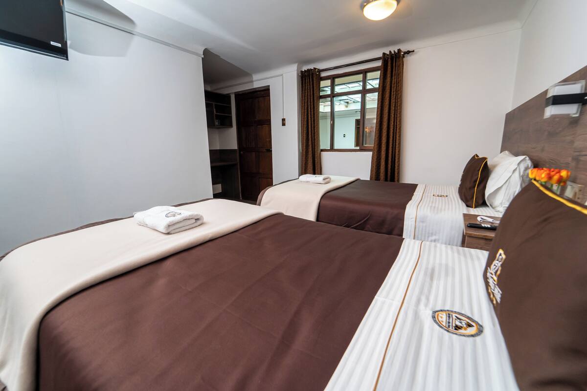 A hotel room with two beds, a wardrobe, a window with curtains, and a bedside table with a lamp and a vase of flowers.