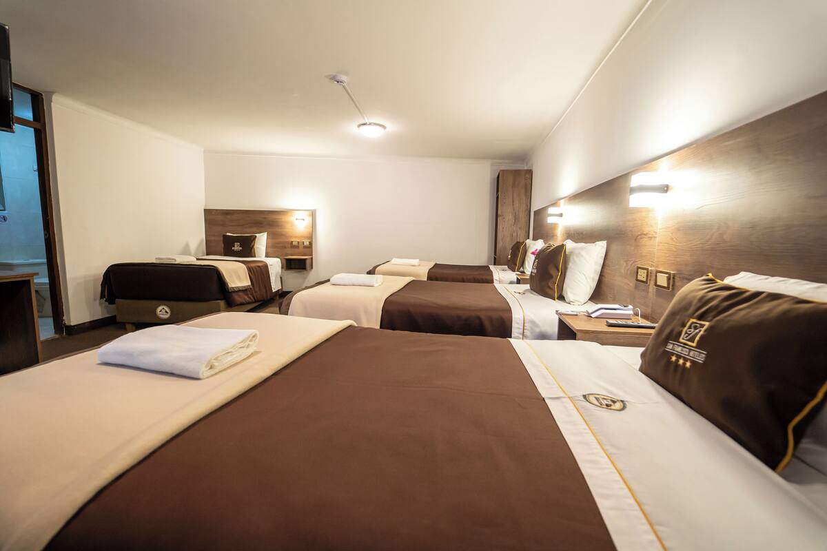 A hotel room with three beds, each with a brown headboard, white linens, and a small bedside table with a lamp.