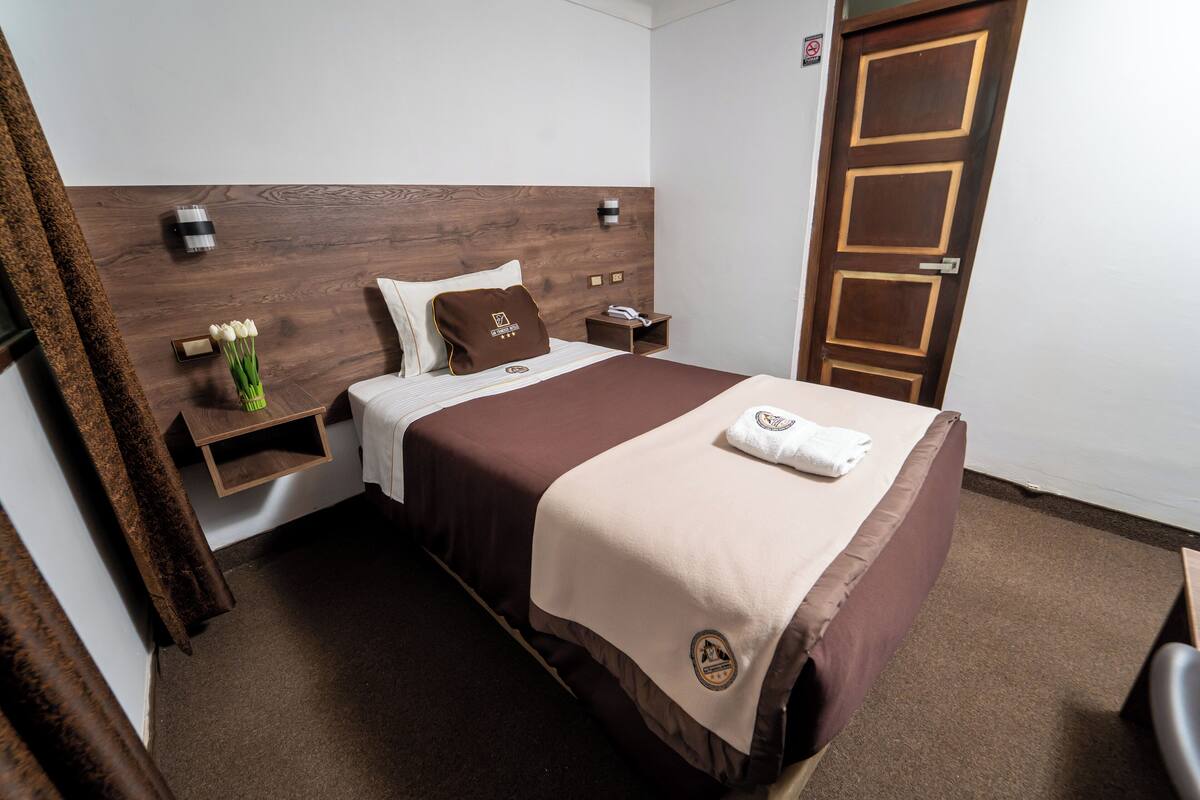 A neatly arranged hotel room with a wooden headboard, a bed with a brown and beige bedspread, a nightstand with a lamp, a small table with a phone, and a door with a wooden panel design.
