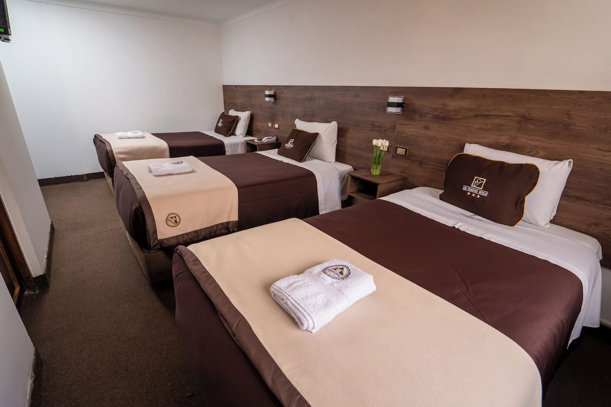 A hotel room with three beds, each with a brown headboard and a beige blanket. There are bedside tables with lamps and a vase with flowers on one of the beds.