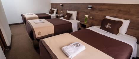 A hotel room with three beds, each with a brown headboard and a beige blanket. There are bedside tables with lamps and a vase with flowers on one of the beds.