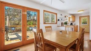 Un comedor amplio con una gran mesa y sillas de madera, una cocina con armarios de madera y ventanas grandes que ofrecen vistas a un área boscosa.