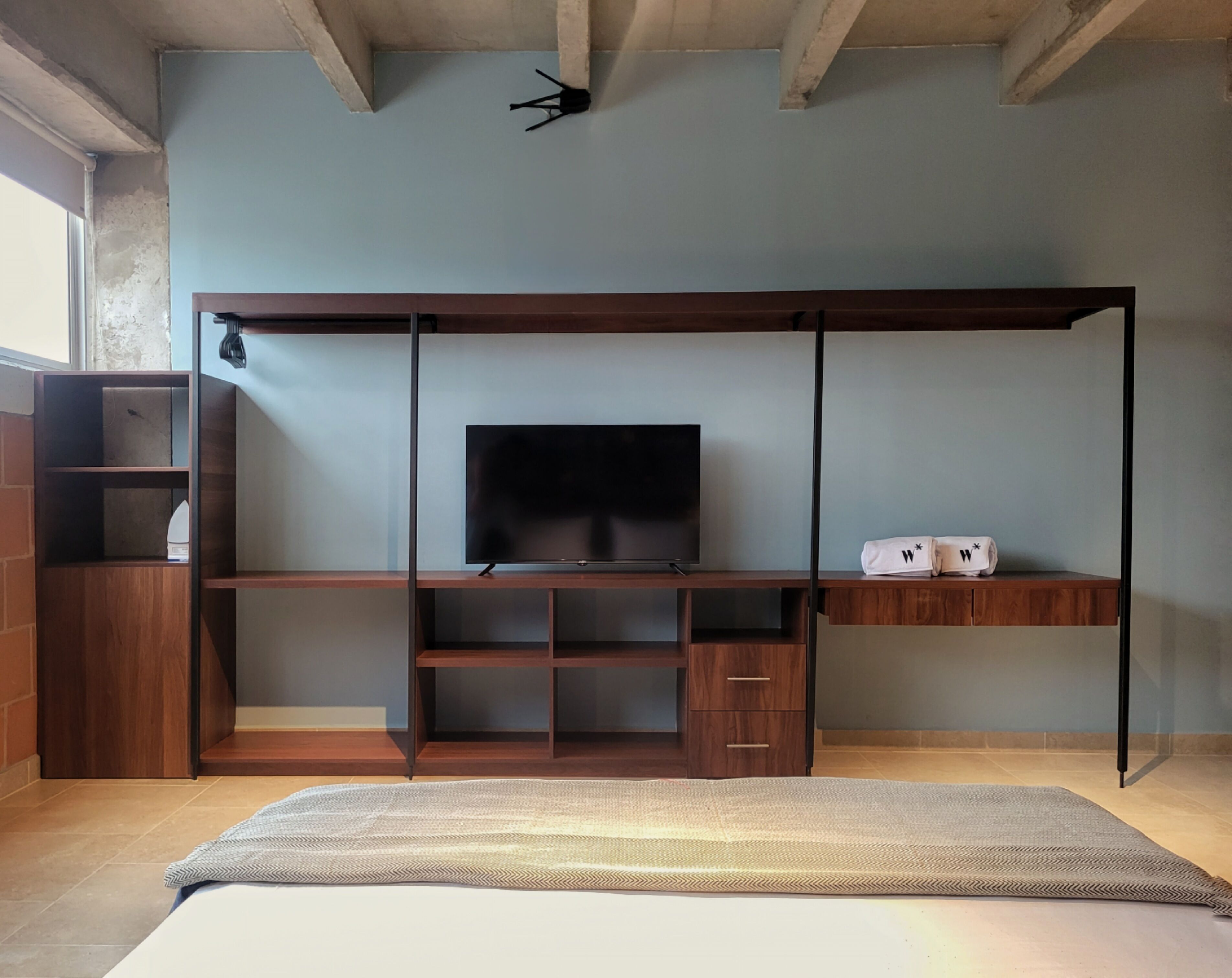 Une pièce comprenant un meuble TV en bois, un téléviseur à écran plat, un petit meuble de rangement et un lit avec un tapis à motifs.