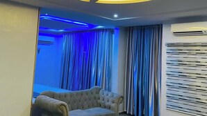 Ein modernes Wohnzimmer mit einer gepolsterten Couch, einer Decke mit runden Leuchten und Blick ins Freie durch ein Fenster mit blauen Vorhängen.
