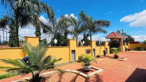 A courtyard with a yellow wall, palm trees, and a covered seating area. - KDP LODGE KIHONDA (Morogoro)