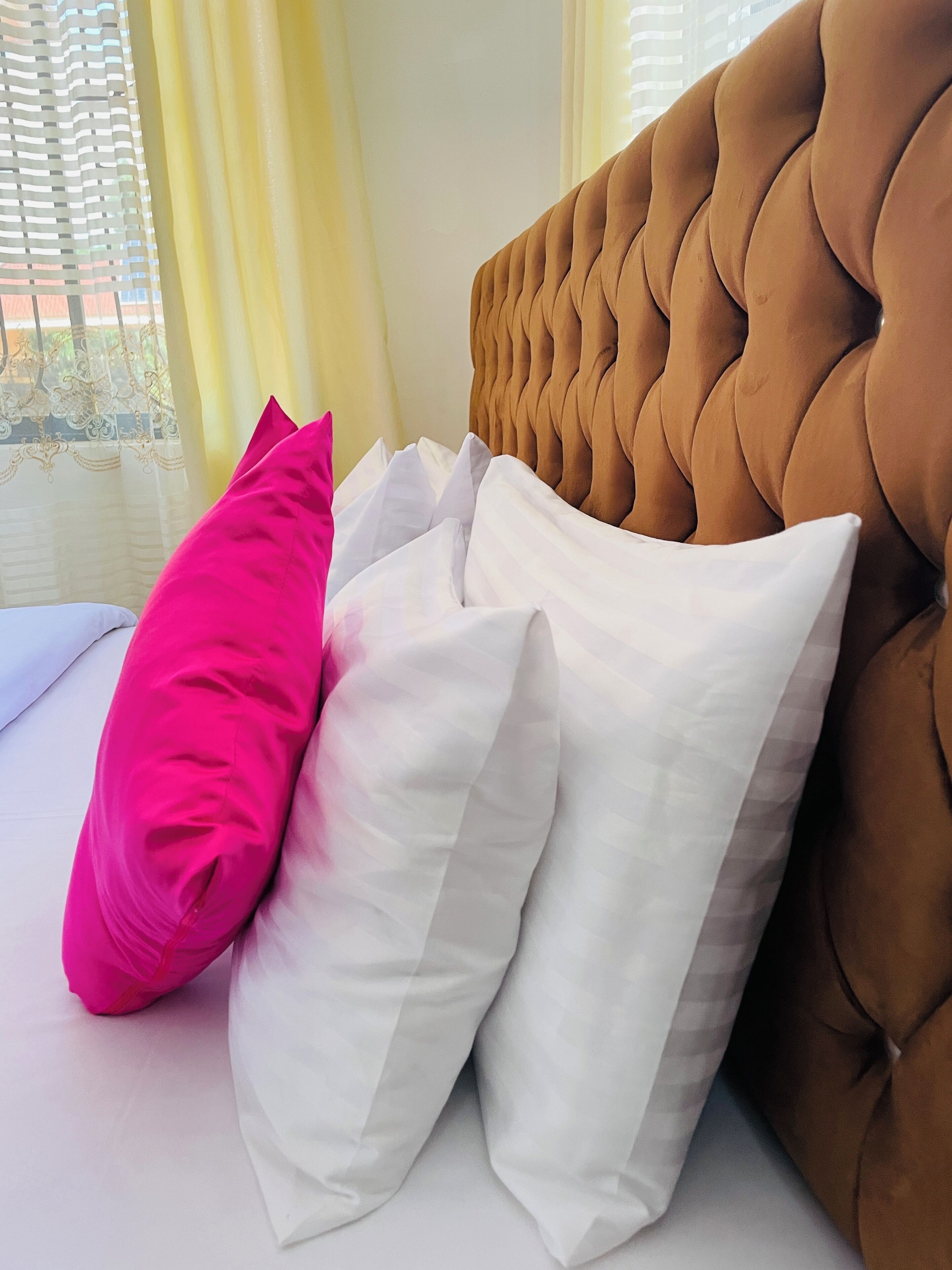 Una cama con cabecera capitonada, almohadas blancas y rosas, y vista al exterior a través de una ventana con cortinas de encaje.