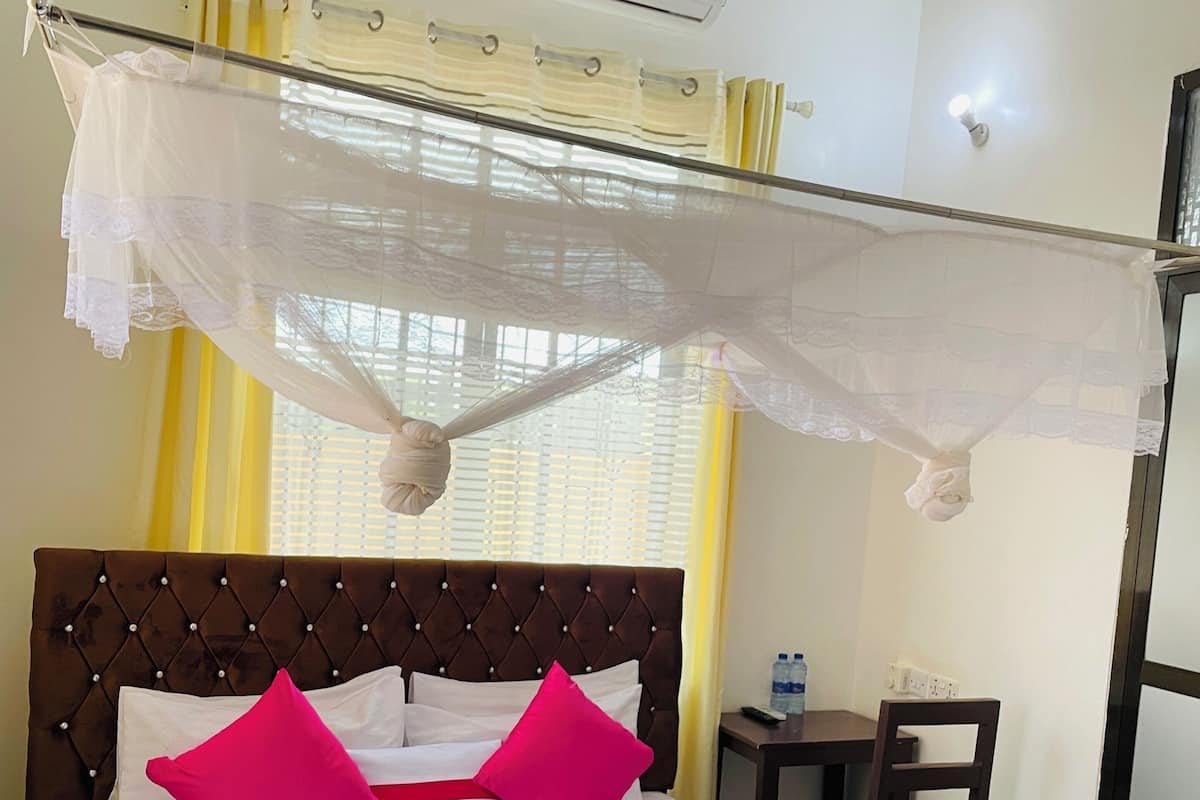 A hotel room with a bed, a bedside table, a chair, an air conditioning unit, and a window with curtains.