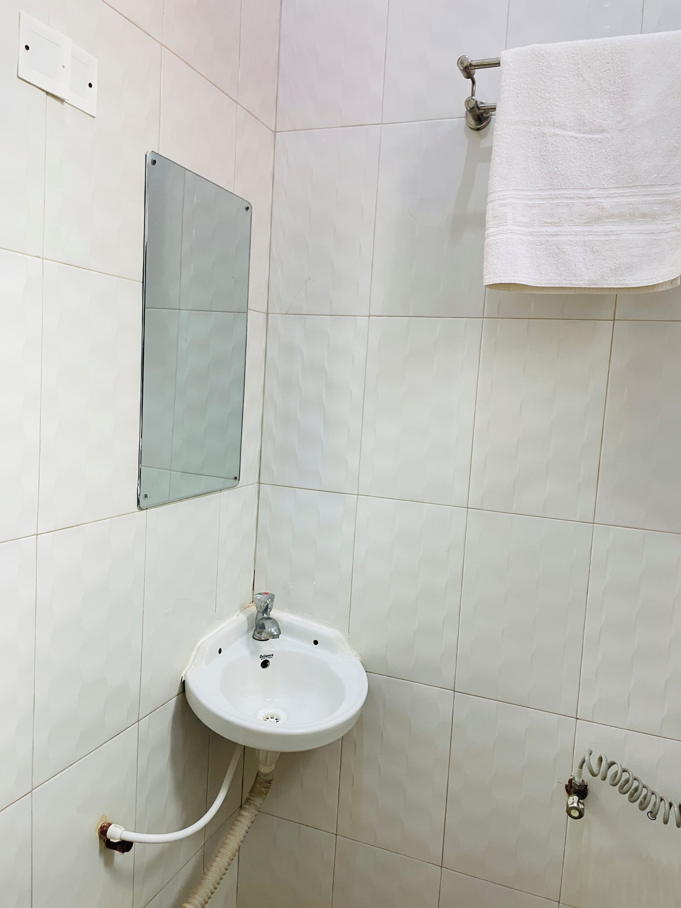 Une salle de bain aux murs recouverts de carreaux blancs, un lavabo blanc avec un seul robinet, une serviette suspendue à un porte-serviettes et un miroir fixé au mur.