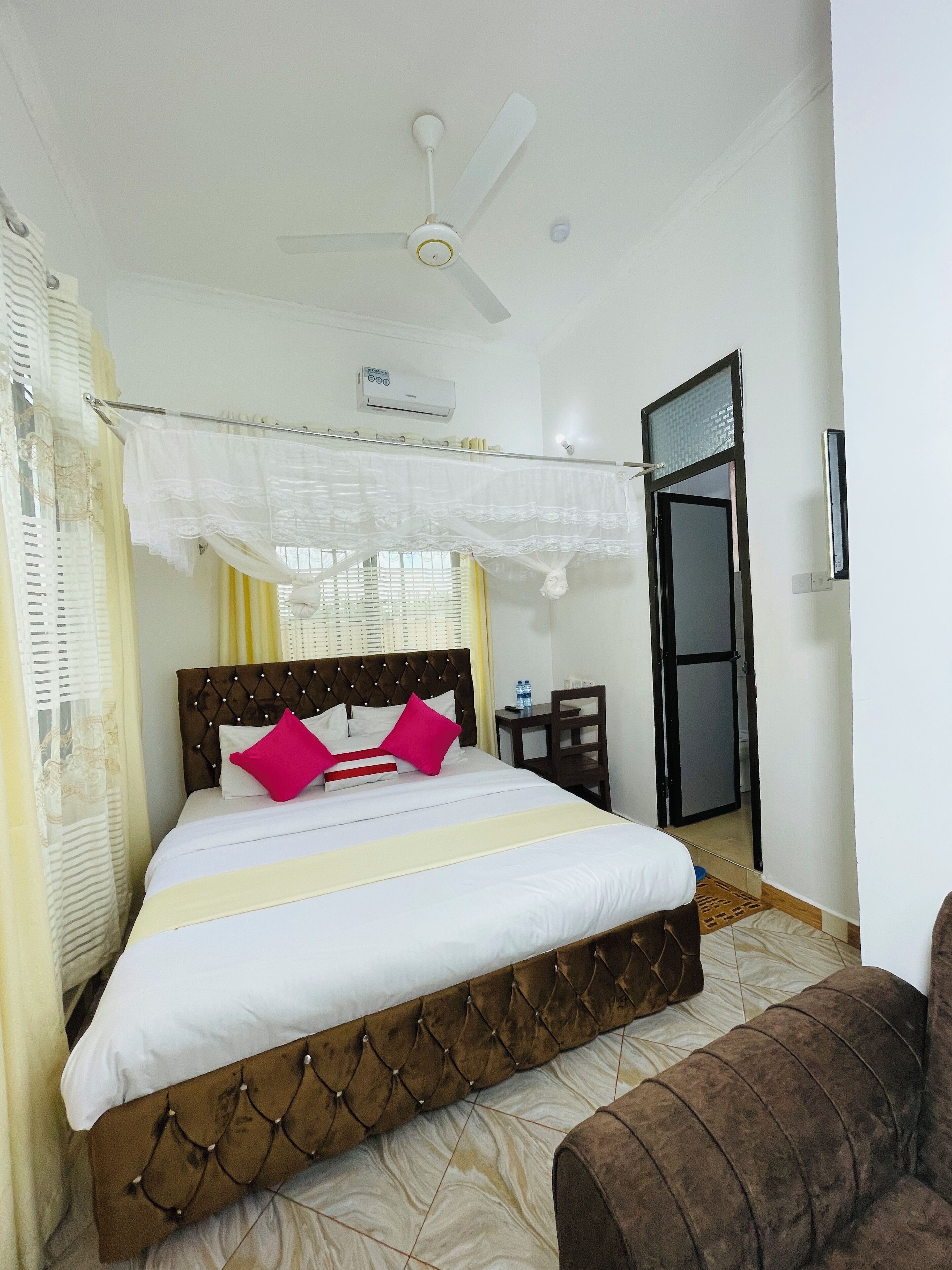 Una habitación de hotel ordenada con una cama, un ventilador de techo, un equipo de aire acondicionado, una mesita con una silla y una puerta que da a otra habitación.
