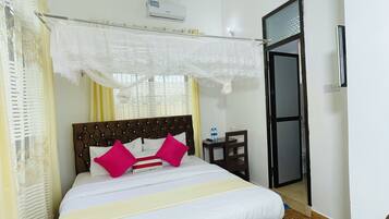Una habitación de hotel ordenada con una cama, un ventilador de techo, un equipo de aire acondicionado, una mesita con una silla y una puerta que da a otra habitación.