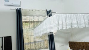 A hotel room with a bed, a ceiling fan, an air conditioning unit, a window with curtains, and a wall-mounted mirror. - KDP LODGE KIHONDA (Morogoro)