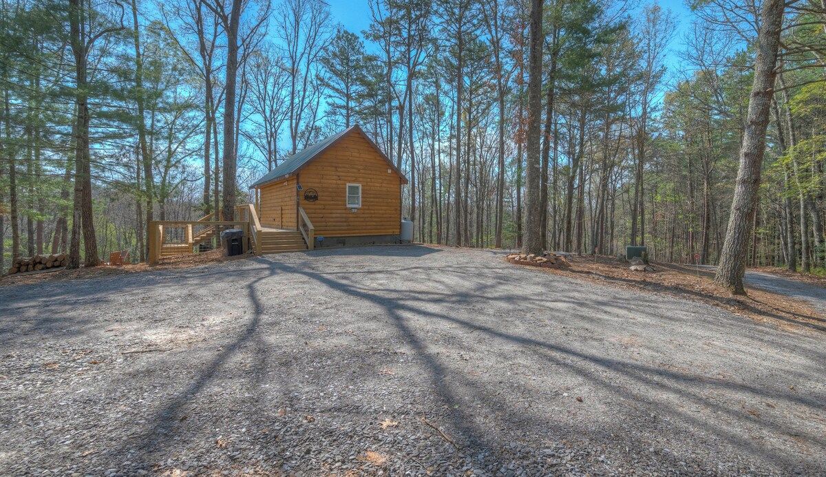 A cabin with a gravel driveway and a wooden deck, surrounded by tall trees.