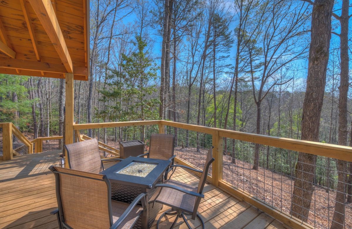 A wooden deck with outdoor furniture and a fire pit, surrounded by a forest.
