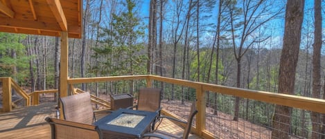 A wooden deck with outdoor furniture and a fire pit, surrounded by a forest.