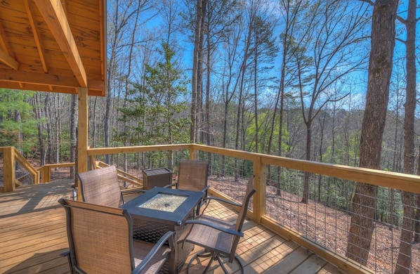 A wooden deck with outdoor furniture and a fire pit, surrounded by a forest.