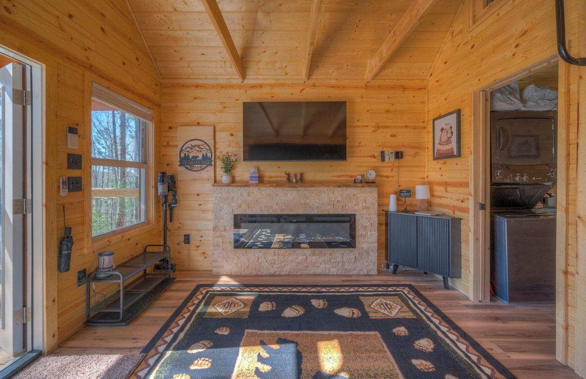 A wooden cabin interior with a fireplace, a television mounted above it, and a kitchen area visible through an open door.