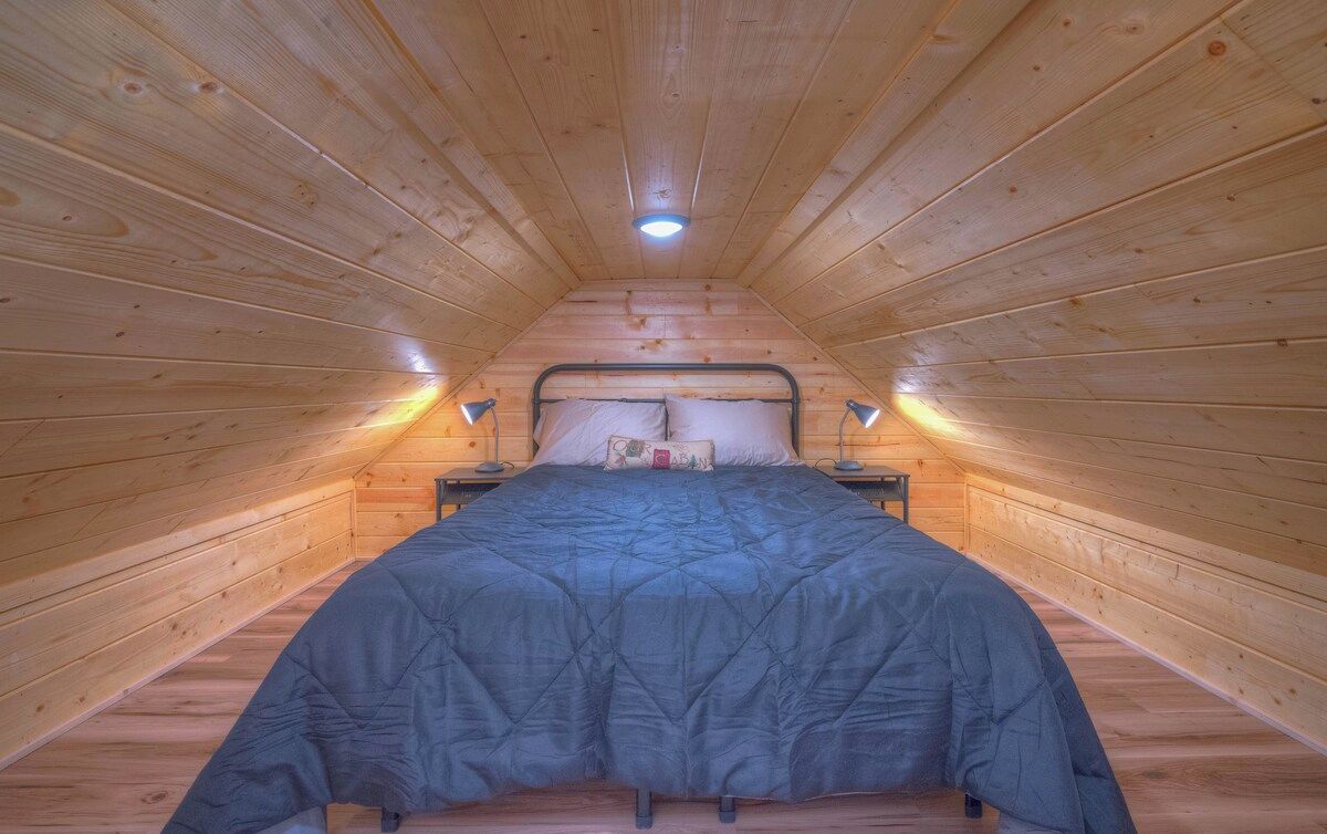 A narrow attic bedroom with a wooden sloped ceiling, a single bed with a blue bedspread, two bedside lamps, and a small nightstand.