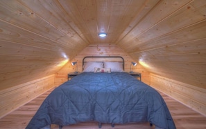 A narrow attic bedroom with a wooden sloped ceiling, a single bed with a blue bedspread, two bedside lamps, and a small nightstand.