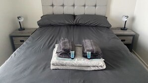A neatly made bed with dark bedding, two bedside tables with lamps, and a folded towel with toiletries on each side.