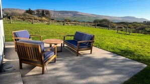 A patio with two wooden chairs and a small round table, overlooking a grassy field and distant hills.