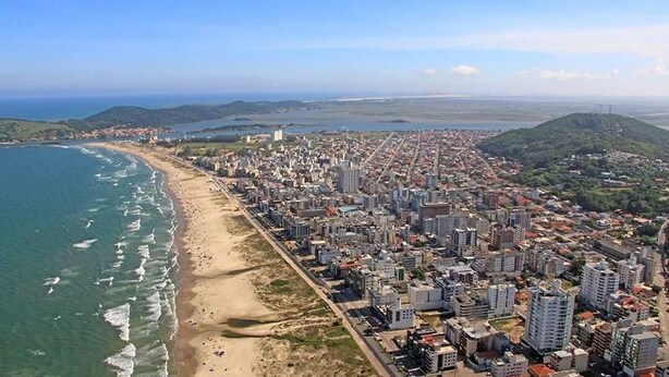 Vista aerea di una città costiera con spiaggia, zona urbana densa e colline sullo sfondo.