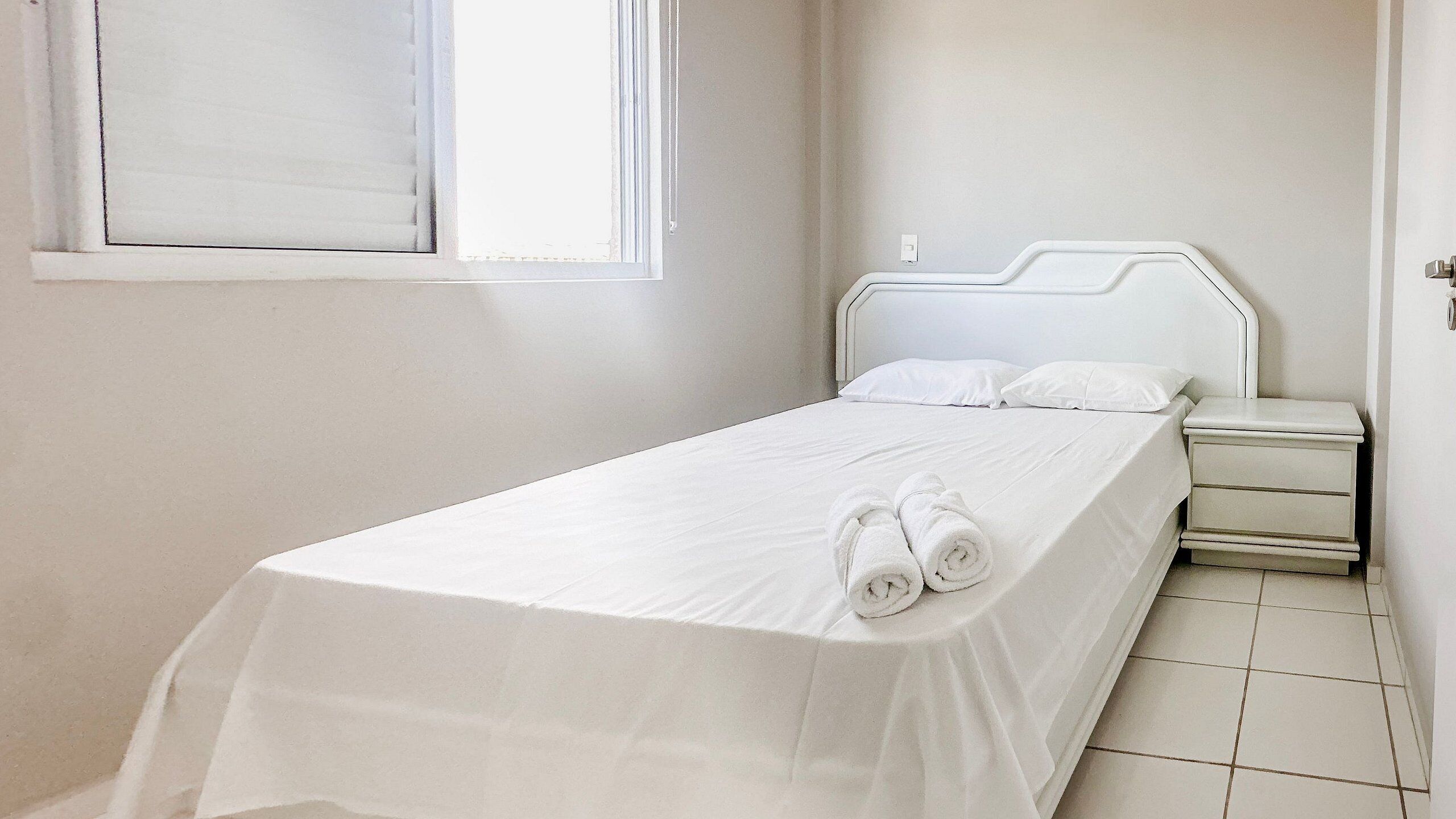 Un lit bien fait, avec du linge de lit et des serviettes blanches, une table de chevet avec une lampe et une fenêtre avec des stores.