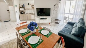 Una cucina moderna con mobili bianchi, un forno a microonde e un frigorifero. C'è una zona pranzo con un tavolo di legno apparecchiato per due, un vaso di fiori blu e un divano nel soggiorno.
